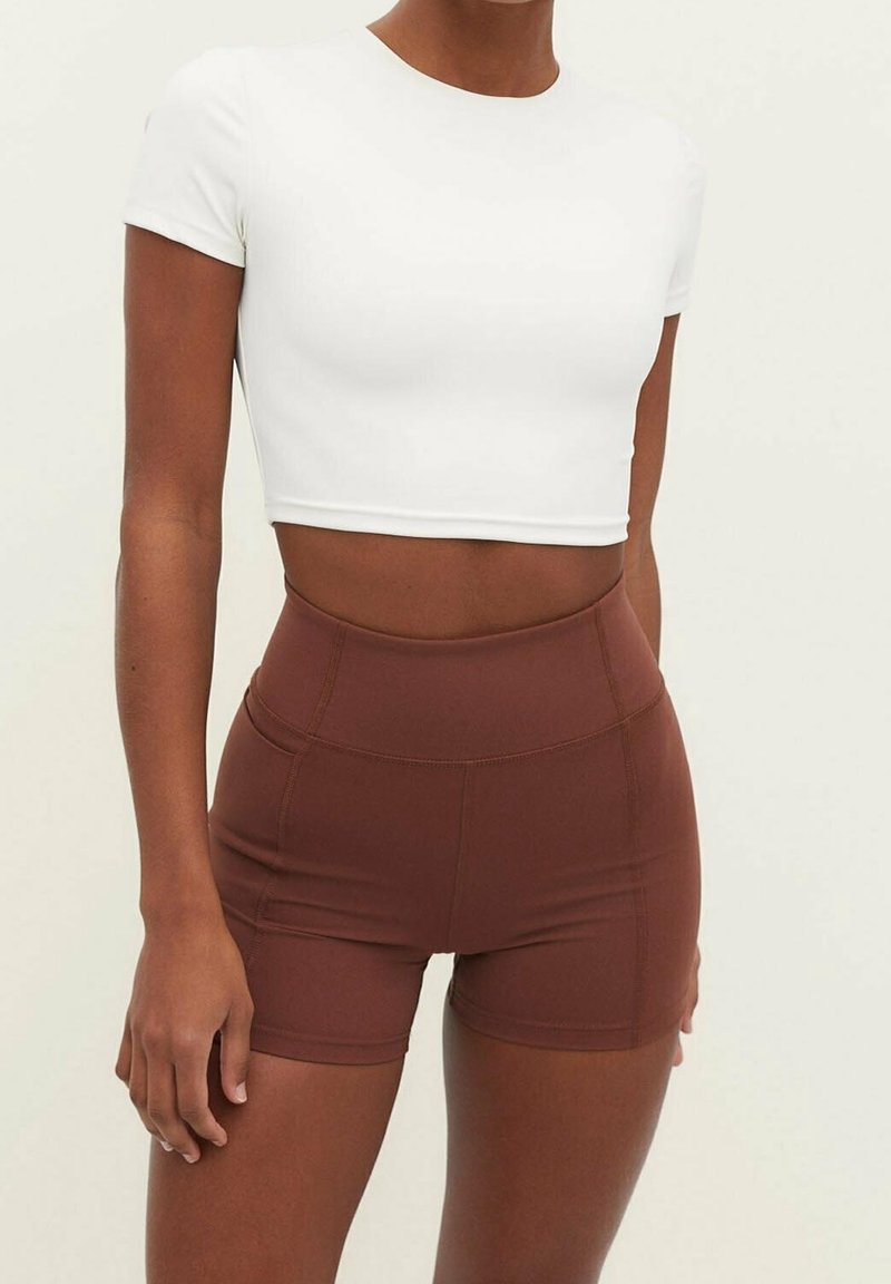 Personne portant un crop top blanc à manches courtes et un short marron cintré taille haute sur un fond uni.