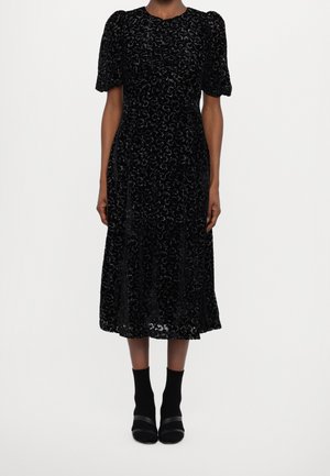 Woman wearing a black patterned midi dress with puffed short sleeves and black ankle boots on a white background.