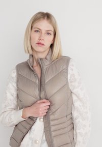 Gilet matelassé beige avec un col haut et une fermeture éclair sur le devant. Présente des coutures diagonales et une coupe ajustée. Porté par-dessus un pull blanc texturé.