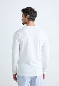 Cămașă albă din bumbac cu mâneci lungi, guler rotund, textură netedă și croială relaxată, purtată împreună cu pantaloni de pijama cu dungi albastre.