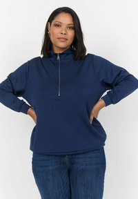 Sudadera azul marino hecha de tejido suave, con cuello de medio zip, mangas largas y un ajuste relajado, combinada con jeans de mezclilla.