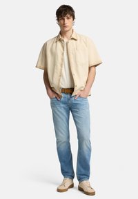 Camicia beige a maniche corte con bottoni sopra una t-shirt bianca, abbinata a jeans azzurri chiari e sneakers beige. Stile casual e vestibilità rilassata.