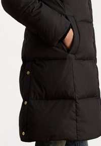 Jachetă puffer neagră cu design quilted, având un buzunar pe lateral, nasturi tip snap și o textură de nailon neted.