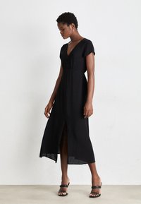 Black maxi dress with short sleeves, V-neckline, and a front slit. Smooth fabric with a fitted bodice and flowy skirt. Black sandals included.