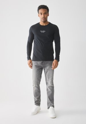 Zwarte langesleeve T-shirt met de tekst "Brooklyn NY", gecombineerd met vervaagde grijze spijkerbroek en witte sneakers, tegen een effen achtergrond.