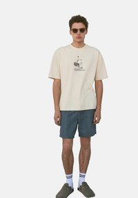 Camiseta de algodón beige con un gráfico de dibujos animados de Snoopy jugando al tenis, combinada con pantalones cortos verde oscuro y zuecos grises de tipo slip-on.