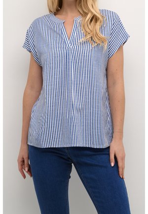 Femme portant un chemisier à manches courtes à rayures verticales bleu et blanc avec un col en V et un jean bleu foncé, debout devant un fond blanc.