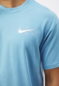 Svetle modré bavlněné tričko s kulatým výstřihem, které má bílé logo Nike a jemný šestiúhelníkový vzor v růžové barvě vedle loga.