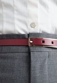 Szürke, szabott nadrág sima textúrával, burgundi bőr övvel, amelynek csillogó arany fém csatja és négyzet alakú kivágásai vannak.