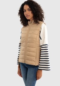 Béžová prošívaná vesta s vysokým límcem a předním zipem; v kombinaci s bílým a námořnickým pruhovaným topem s dlouhým rukávem. Model má na sobě modré džíny.