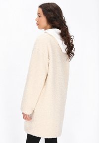 Donna di profilo che indossa un lungo cappotto strutturato color crema sopra una felpa bianca con cappuccio e pantaloni neri, su uno sfondo bianco uniforme.
