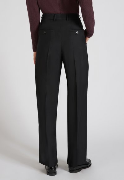 Osoba nosząca czarne klasyczne spodnie z tylnymi kieszeniami i guzikami, w połączeniu z czarnymi butami i ciemnofioletowym swetrem z dzianiny.