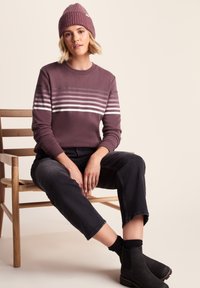 Young woman wearing a mauve striped sweater, matching beanie, black cropped jeans, and black boots, seated on a wooden chair against a beige background.