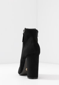 Stivale alla caviglia in suede nero con tacco a blocco, zip posteriore e punta arrotondata, mostrato su uno sfondo bianco.