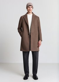 Cappotto marrone in misto lana con taglio sartoriale, caratterizzato da risvolti a lancia, due bottoni e tasche laterali. Abbinato a jeans scuri e scarpe nere.