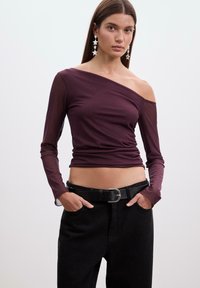 Bordeaux off-shoulder top med lange gennemsigtige ærmer, tætsiddende design og en glat tekstur; båret med sorte højderet pants og et bælte.
