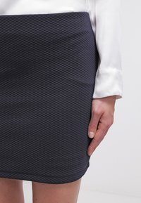 Mörkblå strukturerad kjol med ett diamantmönster, med rak skärning och kant. Tyget verkar tjockt och stadigt. Handen vilar på sidan.