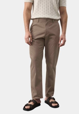 Mand iført beige bukser med lige ben, kremfarvet kortærmet sweater og sorte sandaler med dobbelte remme står mod en hvid baggrund.