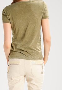 T-shirt verde oliva a maniche corte con una texture morbida, abbinato a pantaloni beige chiaro con tasche posteriori con zip e accenti sottili.