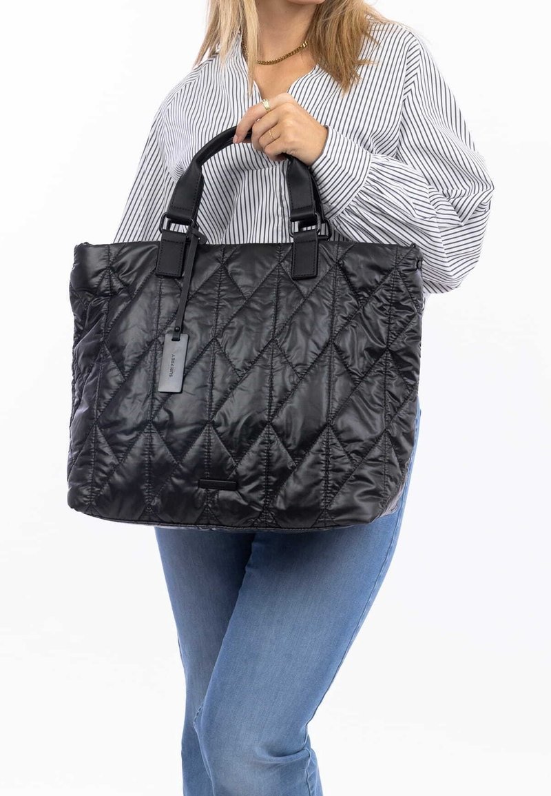Femme portant un jean bleu et une chemise rayée tient un grand sac cabas noir matelassé avec des poignées courtes et une étiquette.