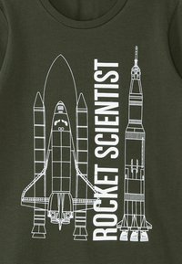 Maglietta verde oliva con disegni a linea bianchi di uno shuttle spaziale e di un razzo, accompagnata dalla scritta "ROCKET SCIENTIST". Collo rotondo.