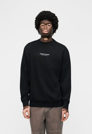 Νεαρός άνδρας με λεύκη που φοράει μαύρο φούτερ με την επιγραφή "WEEKEND OFFENDER ADMIT NOTHING" και γκρι παντελόνι, στέκεται μπροστά σε απλό φόντο.