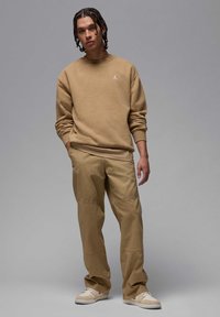 Tan sweatshirt med rund hals og subtilt logo på brystet, parret med matchende tan bukser. Sneakers er lyse beige med hvide detaljer.