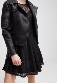 Schwarze Leder-Motorradjacke mit gerippten Schulterakzenten, kombiniert mit einem schwarzen Spitzenkleid mit ausgestelltem Rock und gemusterten Details.