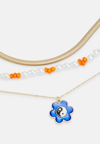 Colar de três camadas com uma corrente de espinha de peixe em ouro, um fio de pérolas com acentos laranja e um pendente de flor azul com um detalhe yin-yang.