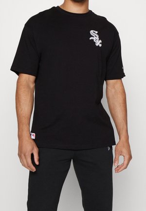 Man draagt een zwart T-shirt met een wit "Sox" logo op de borst en bijpassende zwarte broek, staande tegen een effen achtergrond.