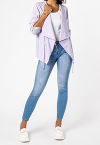 Světlofialový kardigan s překládaným předním dílem a dlouhými rukávy, nošený přes bílý top na ramínka. Modré úzké džíny s lehkým efektem opotřebení a bílé boty.