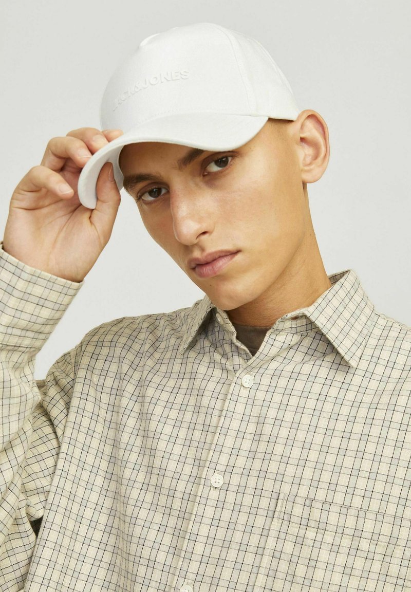 Hvid baseballcap med præget branding, båret i en vinkel. Bæreren har en beige ternet skjorte med knapkrave.