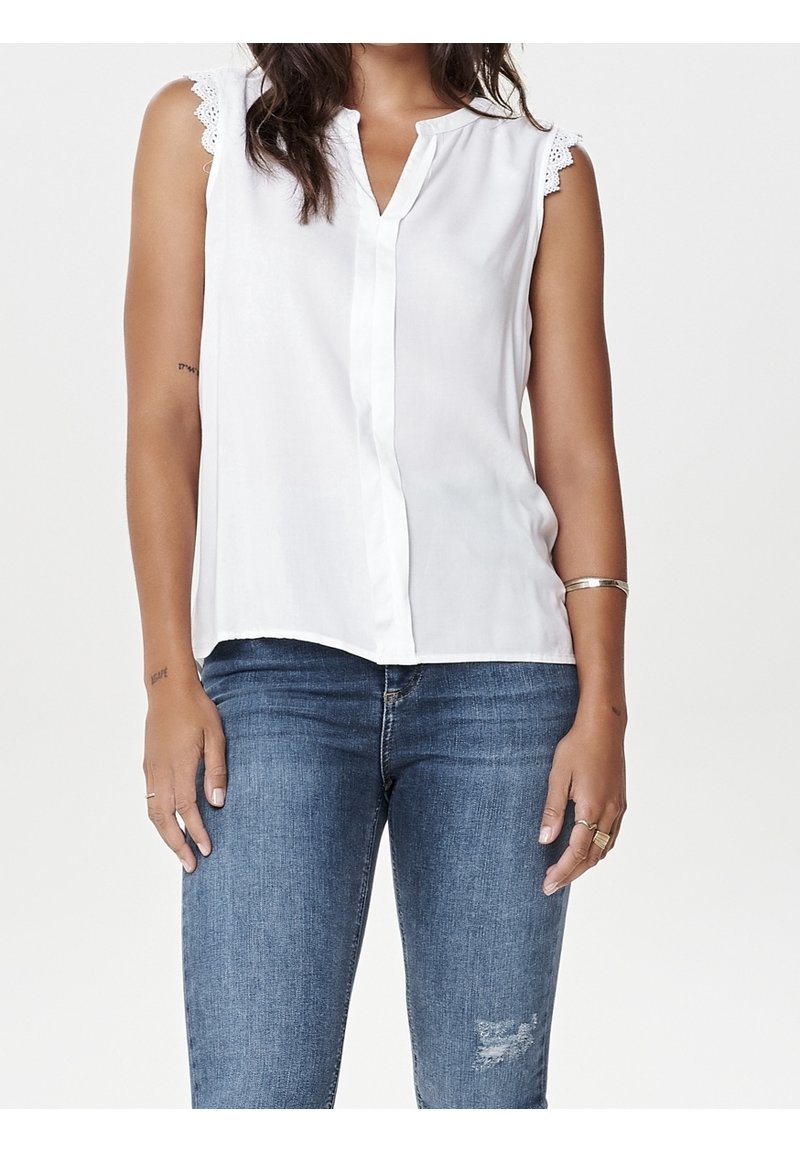 Femme portant un chemisier blanc sans manches avec un bord en dentelle et un jean bleu légèrement usé sur une jambe, debout, les mains relâchées le long du corps.