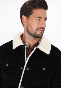 Mann mit kurzem dunklem Haar und Bart, der eine schwarze Cordjacke mit weißem Fleecekragen und ein braunes kariertes Hemd trägt und nach rechts schaut.