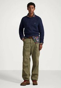 Homem em suéter azul-marinho sobre camisa xadrez vermelha e verde, calças cargo oliva, botas de caminhadas castanhas, em pé com as mãos nos bolsos.