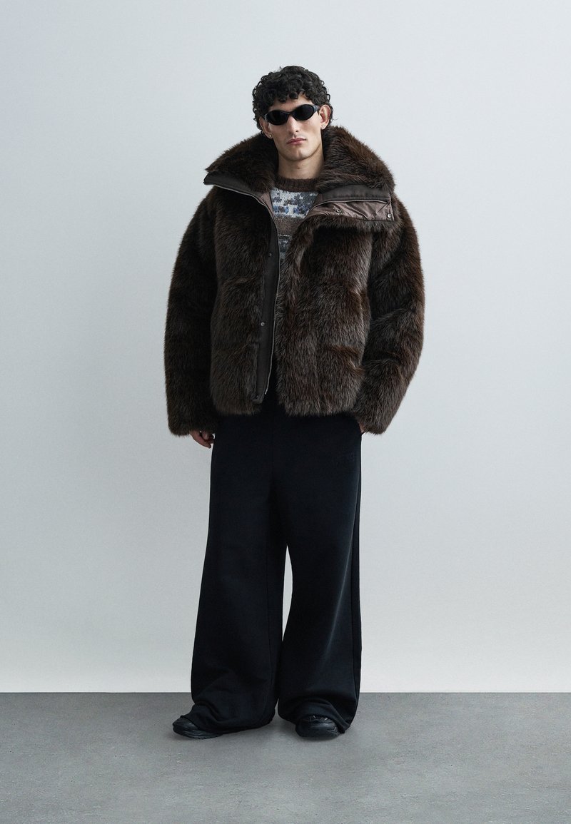 Brown faux fur jacket with a wide collar, front buttons, and a smooth leather panel. Paired with loose black trousers and round sunglasses.