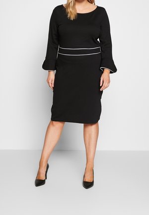 Mujer con un vestido negro hasta la rodilla con mangas acampanadas y detalle en blanco en la cintura, combinado con tacones altos negros.
