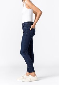Skinny jeans made of dark blue denim, featuring a mid-rise waist, side pockets, and a fitted silhouette, paired with white sneakers.