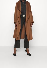 Persona che indossa un lungo cappotto marrone aperto davanti su una camicia di denim nera, jeans neri a vita alta con cintura e scarpe nere con tacco alto.