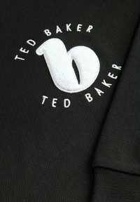 Tessuto nero testurizzato con un logo "Ted Baker" in bianco, ricamato in peluche, e una lettera "b" stilizzata in bianco, circondata da del testo bianco.