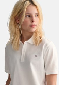 Chemise polo blanche en coton avec un col arrondi, comportant un petit logo brodé sur la poitrine gauche et des manches courtes.