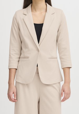 Kvinde iført beige skræddersyet blazer med enkelt knap og matchende bukser over en sort top, stående mod en ensfarvet lys baggrund.