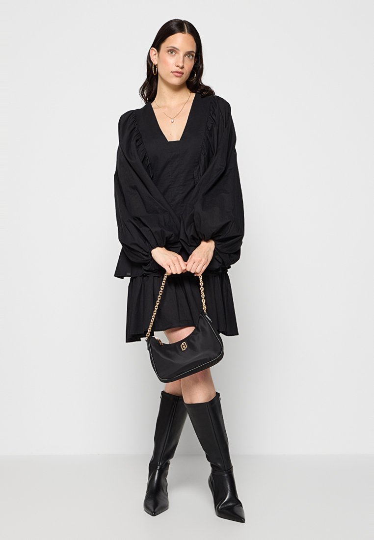 Schwarzes Kleid mit voluminösen Ärmeln und Rüsche, kombiniert mit kniehohen schwarzen Stiefeln und einer kleinen schwarzen Handtasche mit einer goldenen Kettenriemen.
