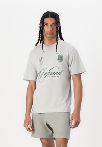 Lys grå t-shirt med kort ærme og grafiske designs i mørkegrøn, med en rund halsudskæring og en afslappet pasform, parret med lysegrønne shorts.
