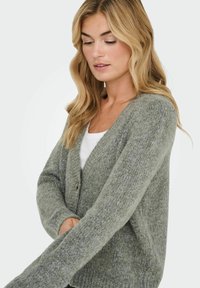 Cárdigan gris de punto con escote en V, cierre de botones y mangas largas. Textura suave con un ajuste ligeramente holgado. Camiseta blanca visible por debajo.