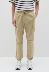 Pantalones cargo beige con cinturilla elástica y cordón. Cuenta con bolsillos laterales y puños elásticos, combinados con zapatillas blancas y grises.