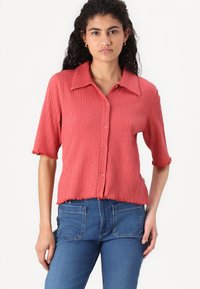 Korte mouwen, koraalrood geribd shirt met een puntkraag, gestructureerde stof, knopen aan de voorkant en gegolfde zoom. Gecombineerd met blauwe jeans.