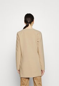 Blazer beige su misura con taglio dritto, tessuto testurizzato e colletto minimalista. La parte posteriore presenta cuciture sulle spalle e una finitura pulita.