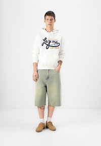 Felpa bianca con stampa blu e gialla, pantaloni shorts in denim verde chiaro, scarpe da ginnastica marroni e calzini bianchi alla caviglia. Design semplice, vestibilità rilassata.