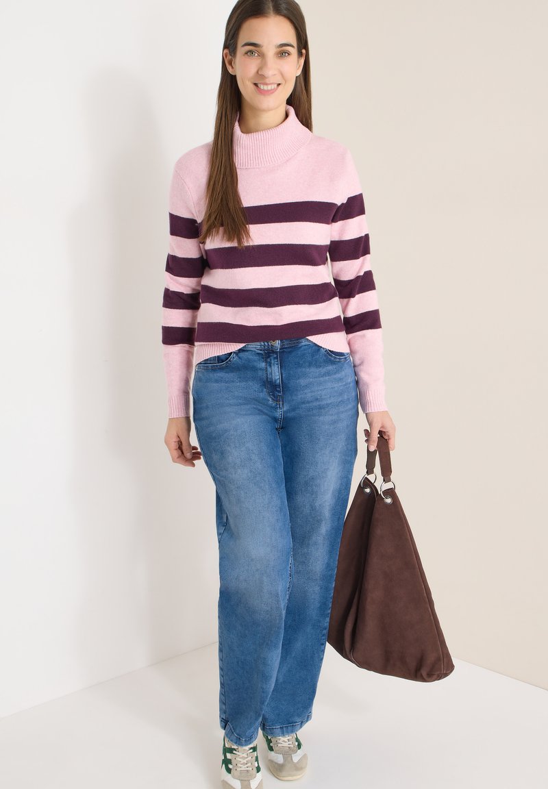 Femme souriante debout contre un mur beige, portant un col roulé rayé rose et violet, un jean bleu, des baskets blanches, et tenant un sac à main marron.